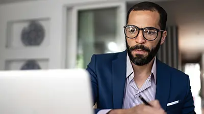 A man wearing glasses and looking at his laptop while taking notes