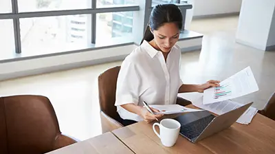 Woman dressed in business casual attire sitting at a table with a coffee mug and laptop while looking over papers.