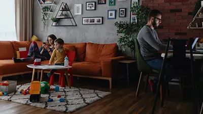 A family in their living room with the father working at his desk while the kids are lounging on the couch. 